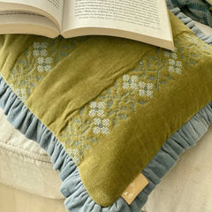 An olive green throw pillow with a blue-gray ruffled border and white embroidered floral patterns, styled with an open book on top.