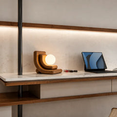 A modern sculptural table lamp with a warm brown wooden base and a glowing white spherical bulb, styled on a white marble-top desk in a minimalist home office setting next to a laptop.