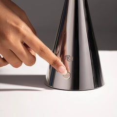 Hand pressing a button on a metallic chrome table lamp with a visible brand logo.