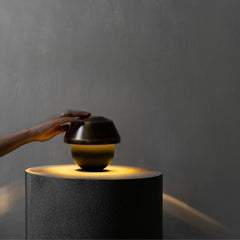 A hand rests on the lid of a small dark metallic tabletop lamp that sits on a cylindrical pedestal, casting a warm yellow light against a textured gray wall.