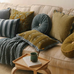 A cozy living room scene with a cream sofa styled with multiple decorative throw pillows in mustard yellow, slate blue, gray, and olive tones, along with a textured gray throw blanket and a small wooden side table holding a green mug and book.