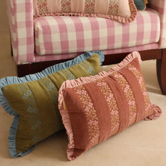 Two decorative throw pillows with ruffled edges displayed on a light beige floor in front of a pink and white checkered sofa. One pillow is olive green with light blue embroidery and a blue ruffle trim, and the other is terracotta pink with gold embroidery and a pink ruffle trim.
