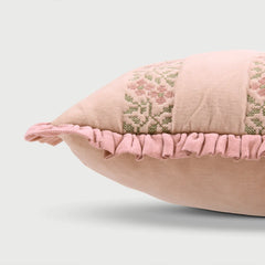 A close-up of a dusty pink decorative throw pillow featuring embroidered floral patterns and a ruffled fabric trim, set against a light gray background.