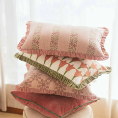 A stack of four decorative throw pillows with ruffled edges, featuring floral and geometric patterns in soft pink, peach, green, and cream tones, styled on a stool in front of a bright window.