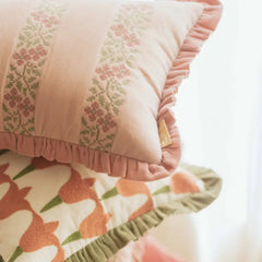A close-up of a soft blush pink pillow with vertical embroidered floral panels and ruffled edges, resting on top of another patterned cushion in a bright, softly lit setting.