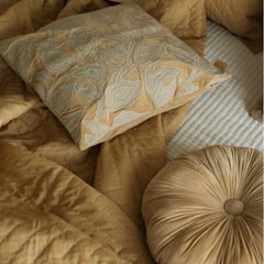 A styled collection of warm neutral home textiles, featuring a tan quilted blanket, a matching round tufted pillow, and a square decorative throw pillow with beige and gray abstract embroidery.