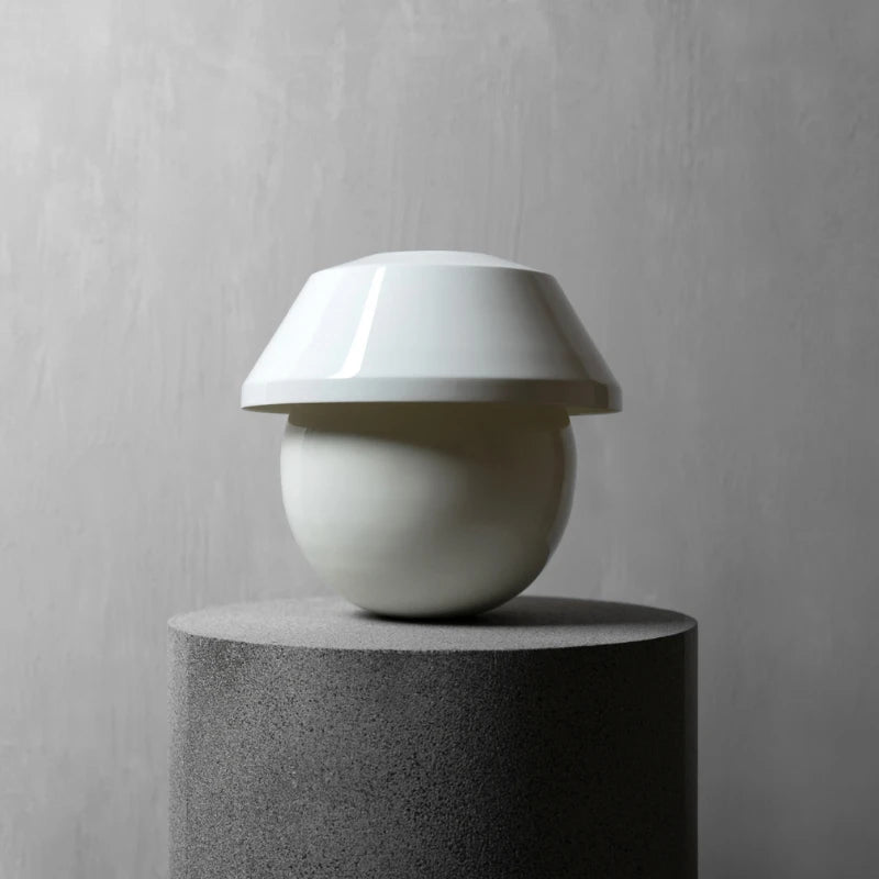 A minimalist white table lamp with a wide, flared shade and a rounded base, displayed on a dark gray cylindrical pedestal against a textured gray wall.