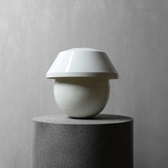 A minimalist white table lamp with a wide, flared shade and a rounded base, displayed on a dark gray cylindrical pedestal against a textured gray wall.