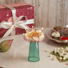 Pushp Glass Diya – teal borosilicate glass stand with blush flower plate and lit diya in jasmine, beside a gift box and pooja thali