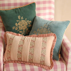 Three decorative throw pillows arranged on a pink and white gingham armchair. The pillows include a dark teal velvet pillow with a gold floral design, a dusty blue velvet pillow with a gold floral design, and a light pink striped pillow with ruffled edges and floral motifs.