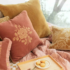 A cozy arrangement of velvet throw pillows in dusty rose and mustard yellow with floral embroidery, styled on a bed with pink patterned bedding and a small tray of cookies.