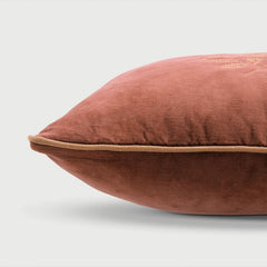A close-up of a dusty rose velvet throw pillow with a beige piping trim, shown against a light gray background.