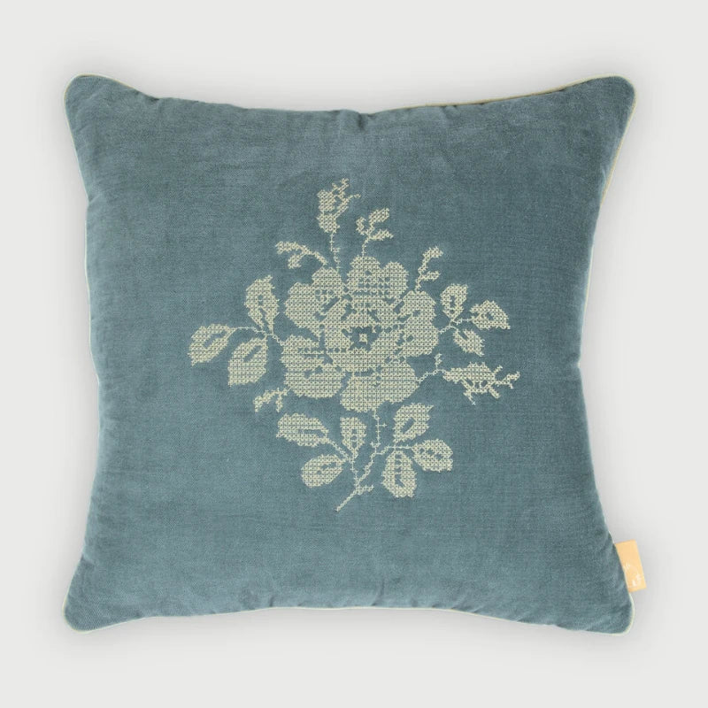 A square teal blue velvet throw pillow featuring a cream-colored embroidered floral motif in the center, with a small yellow fabric tag on the bottom right corner, set against a light gray background.