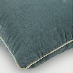 A close-up of a textured teal throw pillow with cream-colored piping along the edge, set against a plain light gray background.