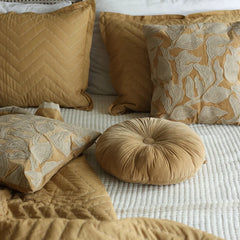 A styled collection of home decor pillows in warm mustard yellow, beige, and cream tones, including a round tufted cushion and patterned pillows on a white textured bedspread.