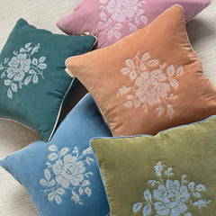 Four decorative square throw pillows in teal, dusty pink, terracotta, and blue-green velvet, each featuring an embroidered floral rose motif in a contrasting color, arranged on a light beige textured surface.