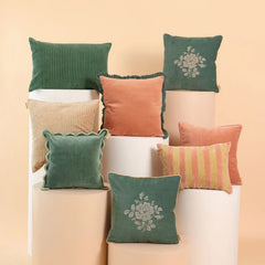 A styled collection of eight decorative throw pillows in various colors and textures, arranged on white cylindrical pedestals against a warm beige background.
