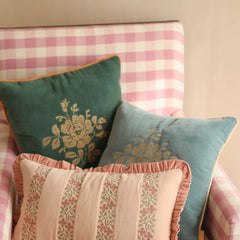Three decorative throw pillows arranged on a pink and white gingham armchair. The pillows include a dark teal velvet pillow with a gold floral design and orange piping, a dusty blue velvet pillow with a gold floral design, and a light pink striped pillow with ruffled trim and floral bands.