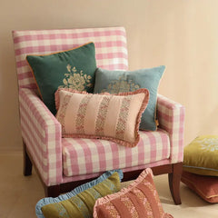 A pink and white gingham armchair styled with multiple decorative throw pillows in green, blue, and pink floral patterns against a warm beige wall.