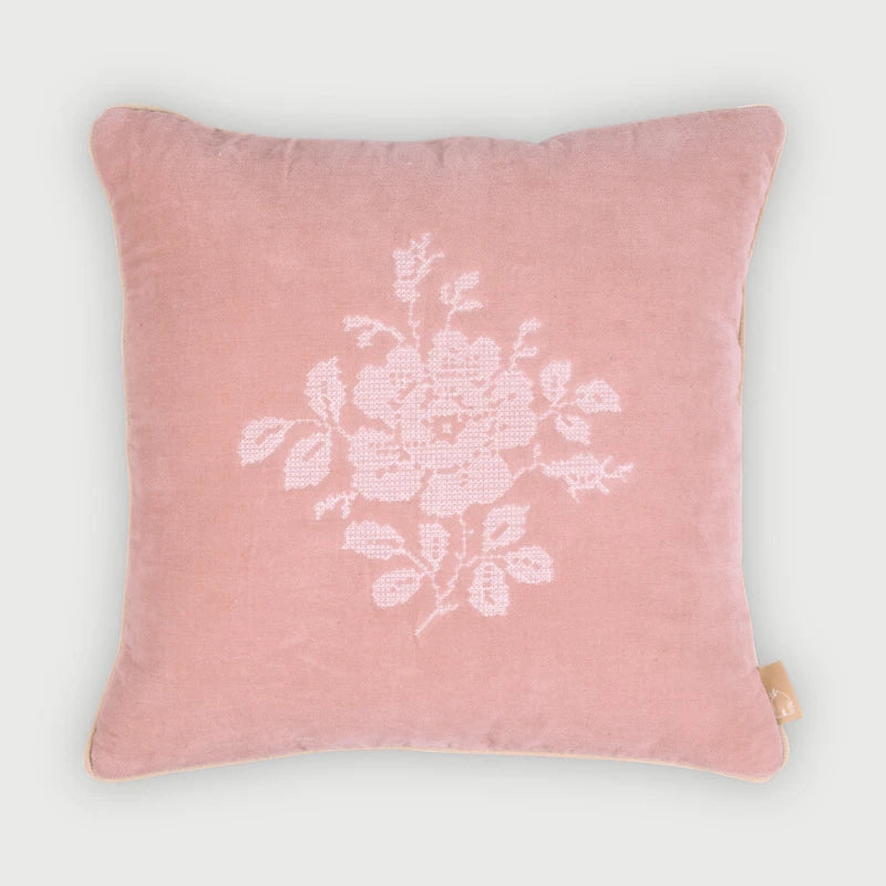 A square pink velvet throw pillow featuring a white embroidered floral motif in the center, set against a clean, light gray background.