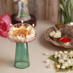 Pushp Glass Diya front view – lit glass diya with jasmine on blush plate and teal stand, pink lotus and pooja thali behind