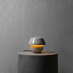 A modern small table lamp with a gray dome shade and a warm glowing spherical base, displayed on a dark gray cylindrical pedestal against a textured gray wall.