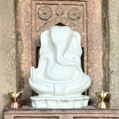 White Polyresin Ganesha Abstract Sculpture of a deity in front of a decorative stone wall with two brass lamps on either side.