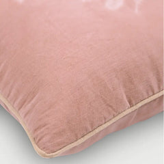 A close-up of a dusty pink velvet throw pillow with a contrasting beige piped edge, shown on a light gray background.