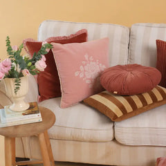 A styled living room scene with a cream striped sofa adorned with multiple decorative throw pillows in dusty pink, rust red, and brown striped fabric, alongside a small wooden side table holding a vase of pink flowers.