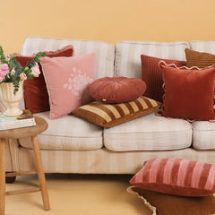 A styled living room scene with a cream striped sofa surrounded by multiple decorative throw pillows in pink, rust red, brown, and striped patterns, plus a small wooden side table with flowers and books.