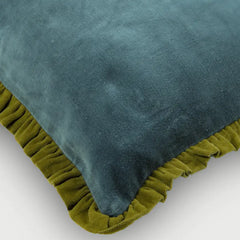 A close-up of a teal velvet throw pillow with an olive green ruffled trim, shown on a light gray background.
