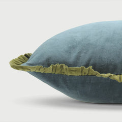 A close-up of a blue-gray velvet throw pillow with a contrasting olive green ruffled trim, set against a clean light gray background.