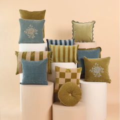 A styled collection of nine decorative throw pillows in various colors and textures, arranged on white cylindrical pedestals against a solid pale peach background.