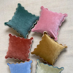 Six velvet throw pillows in assorted colors including teal, pink, terracotta, mustard, light blue, and sage green, arranged on a light beige textured surface.