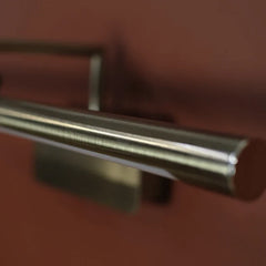 Brass Antique Picture Light, close-up of a brushed metal cylindrical handle or fixture mounted against a warm reddish-brown background.