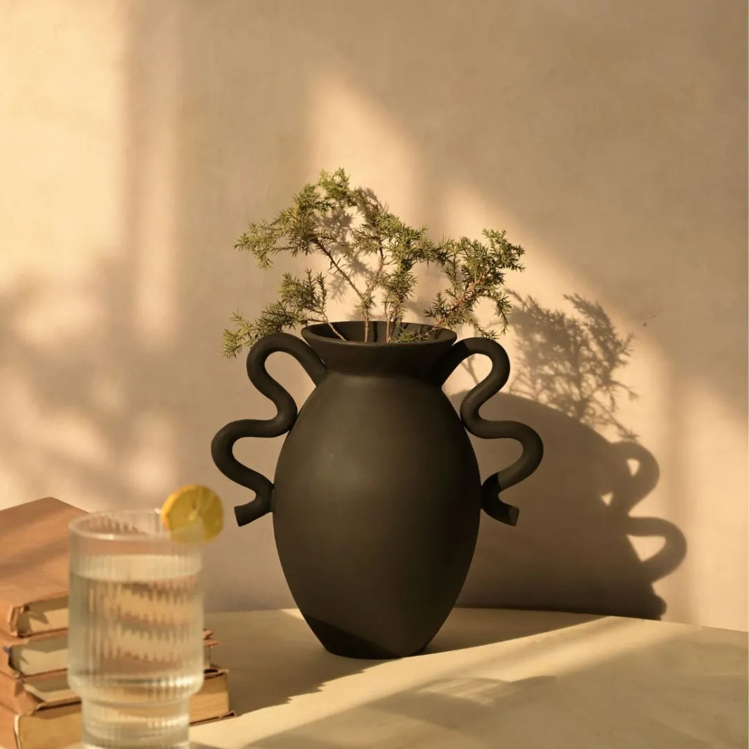 Black Asteria Vase with plant on a table next to a glass of water and lemon slice, soft lighting