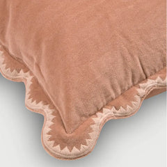 A close-up of a dusty rose velvet throw pillow with a scalloped edge and embroidered detailing, set against a light gray background.