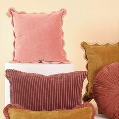 A styled collection of velvet and corduroy throw pillows in pink, rust, and mustard yellow, arranged on a white pedestal against a warm beige background.
