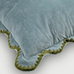 A close-up of a dusty blue velvet throw pillow with a scalloped edge trimmed in olive green embroidery, shown on a light gray background.