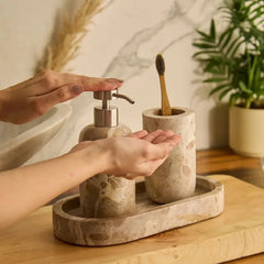 A bathroom sink toiletry set made of beige stone with a Koru Beige soap dispenser and toothbrush holder, shown with hands using the dispenser and a wooden toothbrush in a real bathroom setting.