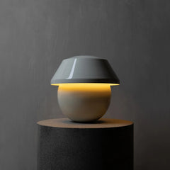 A modern table lamp with a glossy dark gray dome-shaped shade and a matte beige spherical base, illuminated with a warm yellow glow from within. The lamp is displayed on a dark cylindrical pedestal against a textured gray wall.