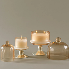Sundance Aura Candle, small and medium size candles. Three lit candles in glass holders with matching glass cloches, arranged on a neutral gray background.
