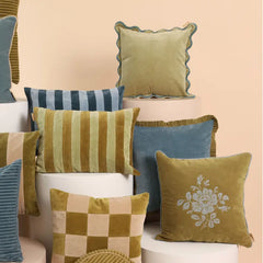 A collection of decorative throw pillows in muted olive green, blue, beige, and striped patterns, styled on white cylindrical pedestals against a soft peach background.