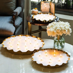 Three scalloped gold metal Flower Tray Candles in Damask Rose filled with lit white pillar candles are arranged on a table in a living room setting, with a vase of white roses and a matching set on a side table in the background.