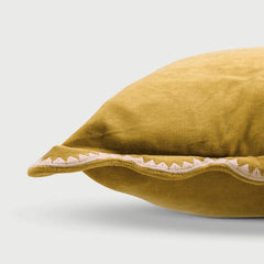 A close-up of a mustard yellow velvet pillow with a scalloped edge and white embroidered detailing, set against a light gray background.