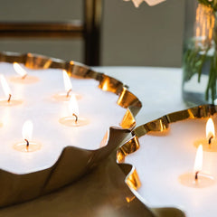 A close-up of a Flower Tray Candle in Damask Rose fragrance and in a scalloped metallic gold petal-edged rim tray, lit with several small tealight candles on a tabletop.