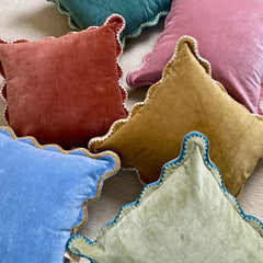 A group of velvet throw pillows in assorted colors including blue, green, rust red, mustard yellow, and dusty pink, arranged on a light beige textured fabric background.