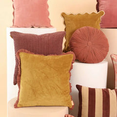 A styled collection of velvet and corduroy throw pillows in dusty pink, mustard yellow, and rust red, displayed on white cylindrical pedestals against a warm beige background.