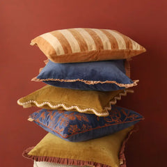 A stack of five decorative throw pillows in various colors and textures, including striped velvet, ruffled velvet, and embroidered fabric, set against a solid dark reddish-brown background.