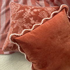 Two decorative throw pillows in a warm rust-orange velvet fabric, styled on a striped bedspread. The front pillow is solid rust-orange with a scalloped edge, while the back pillow features a lighter floral pattern on a slightly darker rust-orange background.